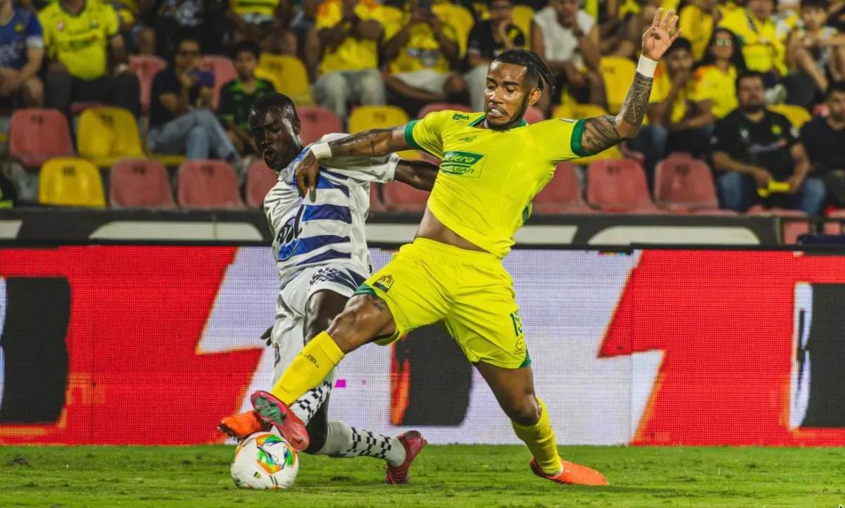 Jugador del Atlético Bucaramanga con uniforme amarillo disputa el balón ante un rival en un partido de la Liga BetPlay, con hinchas auriverdes en la tribuna